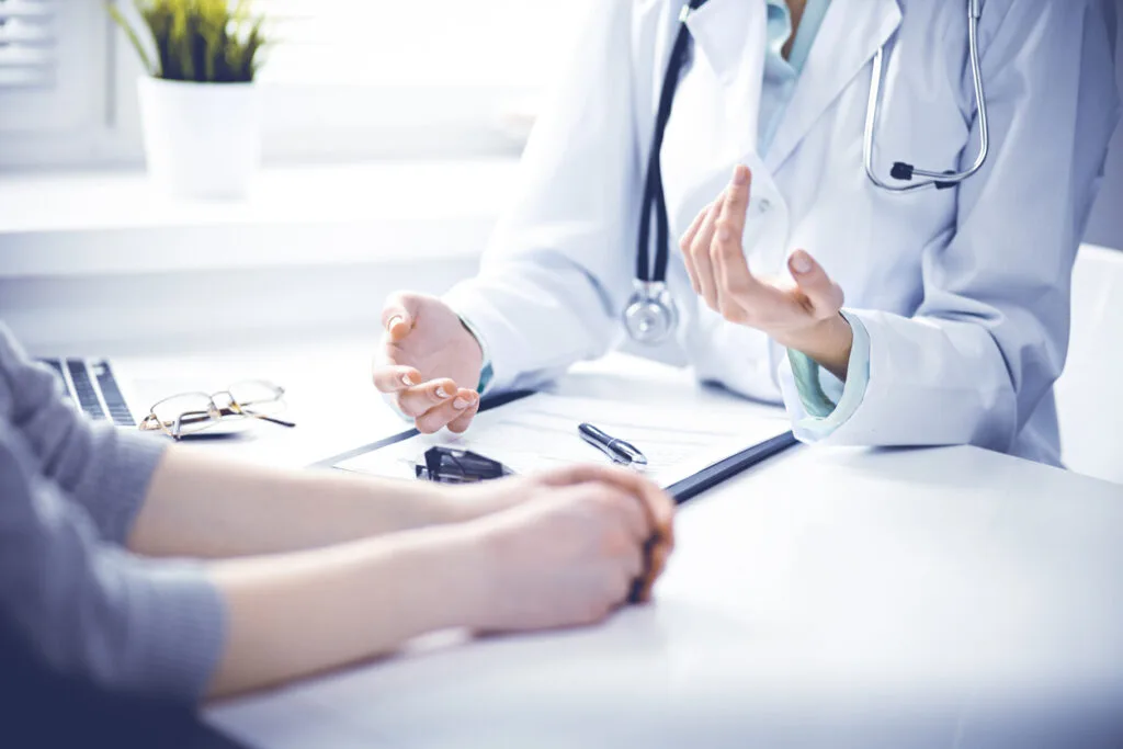 Doctor and female patient sitting at the desk and talking in clinic near window.